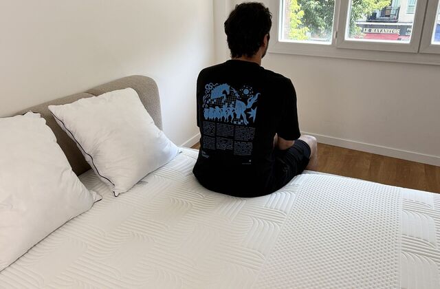 Homme assis de dos au bord d'un matelas, face à la fenêtre