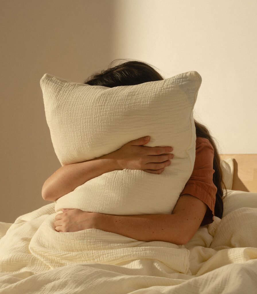 Oreiller Universel Tediber dans une taie claire, tenu dans les bras sur un lit
